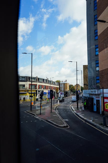 View of a city street through a narrow window or opening, showing a curved road with double yellow lines, traffic moving in both directions, and pedestrian crossings. On the left side, there is a row of commercial buildings, including a shop with a yellow and black sign, while above, a bus and several cars are visible. The sky is blue with scattered white clouds, and streetlights line the road. The scene depicts an urban environment typical of a residential and commercial area in Finsbury, with nearby high-rise buildings on the right. As part of home relocation or furniture transport processes, this image captures the exterior environment and street layout relevant to the logistical planning for a move, possibly illustrating the approach to a property where household items are being loaded or unloaded. Man with Van Finsbury, a professional removals company, might utilize such scenes in their service overview for same-day moving options from Finsbury Park to Old Street.