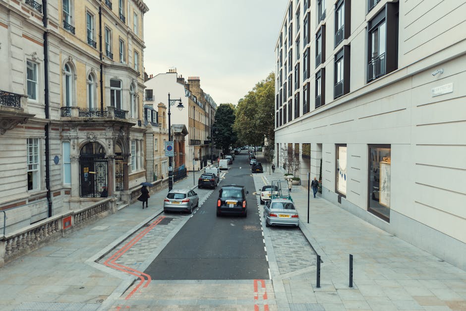 A view of a city street taken from an upper floor or window, showing parked cars along the pavement and buildings on both sides. On the left, there is a historic-style building with ornate architectural details and multiple windows, while on the right, a modern building with a smooth, light-colored facade and large windows is visible. Pedestrians are walking along the sidewalks, and a person holding an umbrella is seen near the building on the left. The street appears to be used for parking, with several vehicles, including a black van parked close to the curb. The overall scene suggests an urban environment suitable for house removals or furniture transport, in line with services provided by Man with Van Finsbury during home relocation or moving operations, as indicated on the page about handling awkward items in City Road terraces, FINSBURY.