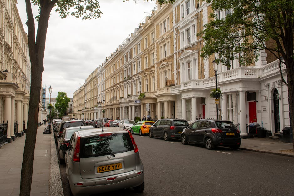 A row of white Victorian terraced houses along City Road in Finsbury with large bay windows and decorative mouldings. The street is lined with parked cars, including a silver van, several small hatchbacks, and a yellow sports car, parked on both sides of the road. A few trees are visible along the sidewalk, with green foliage partially shading the pavement. In the foreground, a man with a move trolley is seen carrying a large, wrapped piece of furniture towards the entrance of one of the terraced houses. The scene is set during daylight under cloudy skies, with no visible moving trucks but indications of ongoing home relocation activities in the area. The image focuses on the typical urban street scene relevant to house removals and furniture transport services provided by Man with Van Finsbury, highlighting the process of handling and loading awkward or large items during a residential move.