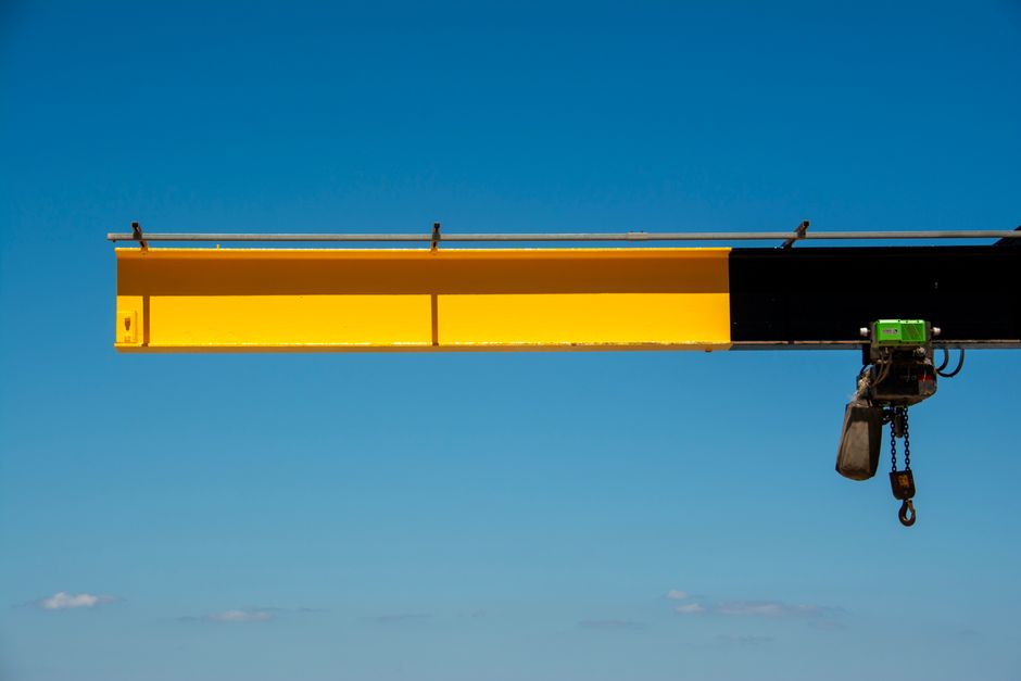 A large, yellow overhead crane arm extending horizontally against a clear blue sky, with a black section on its right side containing a small green motorized pulley system and a hanging chain hoist. The crane is part of a vehicle or loading equipment used in house removals or furniture transport, visible in a moving or loading context similar to what is described in the page about parking suspensions and hoists with Islington Council. The crane's structure is used to lift heavy furniture or boxes during the home relocation process managed by Man with Van Finsbury, with the equipment positioned above a loading area or external driveway for efficient packing and moving activities.