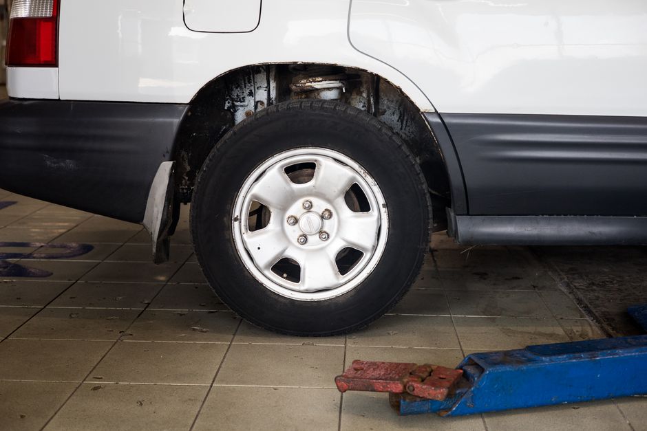 A close-up view of the rear wheel of a white commercial van parked indoors on a tiled floor. The wheel has a white steel rim with five large, rounded cutouts and is fitted with a black tire. The van's wheel arch shows some dirt and rust, indicating usage and exposure to the elements. Part of the vehicle's black side trim and lower panel is visible beneath the sliding door. In the foreground, a blue hydraulic wheel dolly with a red padded grip is positioned near the van, suggesting a loading or moving process. The indoor environment appears to be a garage or vehicle servicing area with even lighting, clean tiled flooring, and no other objects or furniture in sight. This image illustrates a typical scene during home relocations involving furniture transport and vehicle loading with professional moving equipment, as provided by Man with Van Finsbury.