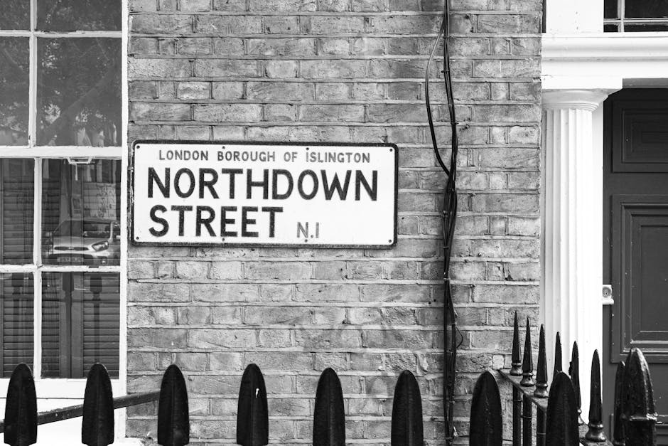 A black-and-white photograph of a residential exterior showing a brick wall with a London Borough of Islington street sign indicating 'Northdown Street, N1', affixed to the wall just above a black iron fence with pointed finials. To the left of the sign, there is a window with multiple panes, reflecting parked cars and some interior furnishings. On the right side, there is a white column featuring classic architectural detailing, supporting a porch or entrance area. A black cable runs vertically along the brick wall, secured at intervals with clips. The scene is well-lit, with natural light highlighting the textures of the brickwork, the metal fence, and the architectural features, capturing details relevant to home relocations, furniture transport, or moving logistics for a property situated on Northdown Street.
