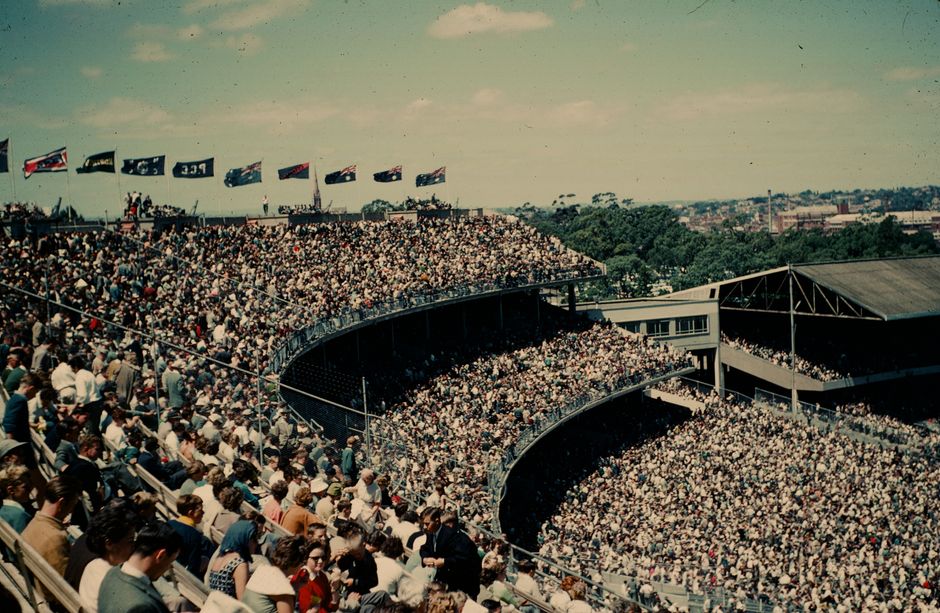 A large outdoor stadium filled with a densely packed crowd of spectators seated in curved tiered rows, with some individuals standing and walking along the aisles. The crowd consists of people wearing various casual clothing, including jackets, hats, and dresses. In the background, there are multiple flags flying on flagpoles, including what appears to be national or team banners, positioned above the crowd. The stadium structure includes a metal fence barrier along the front edge of the seating area, and a section of the field or stage is not visible in this view. Surrounding the stadium, there are trees and distant buildings under a clear, slightly cloudy sky, indicating daytime. The scene captures the busy atmosphere of a sports event or large gathering, with some movement and activity typical of home relocation or transport movements within a stadium setting, relevant to understanding logistics in moving processes as reflected in the context of house removals and transport services.