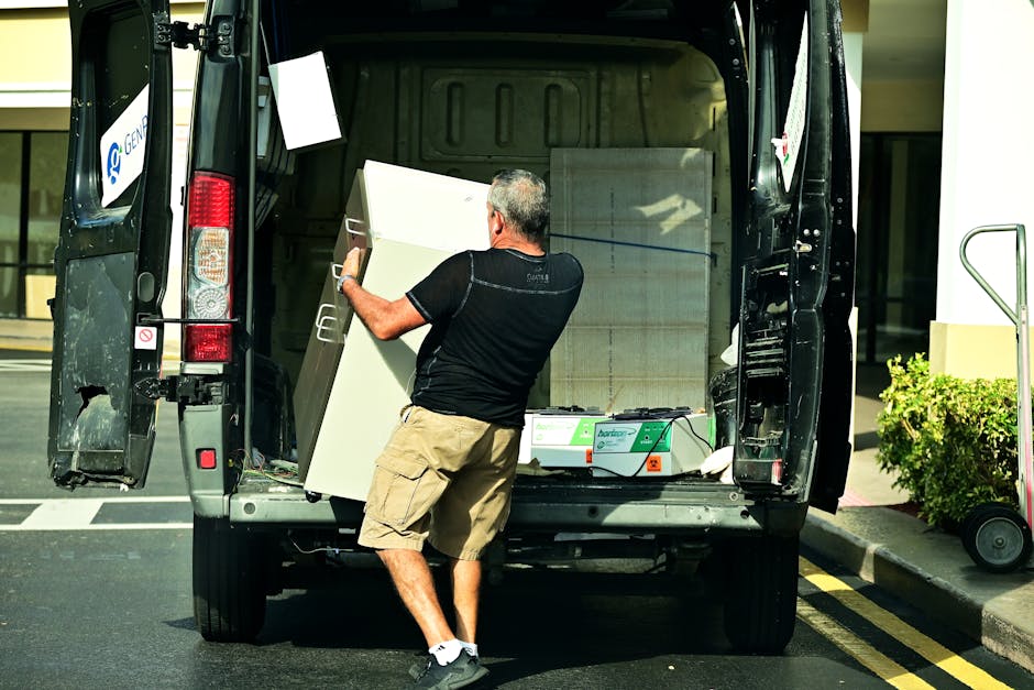 A man in casual clothing, wearing a black t-shirt and beige shorts, is loading a large white kitchen cabinet into the rear of a black moving van parked on a street. The van's back doors are open, revealing the interior where other packed boxes and packing materials are stored. The cabinet is being lifted from the ground and positioned inside the vehicle, with a hand gripping the side for stability. The surrounding environment includes a sidewalk with a potted shrub, and the scene appears to be part of a home relocation or furniture transport process operated by Man with Van Finsbury, as indicated by the company's branding on the van.