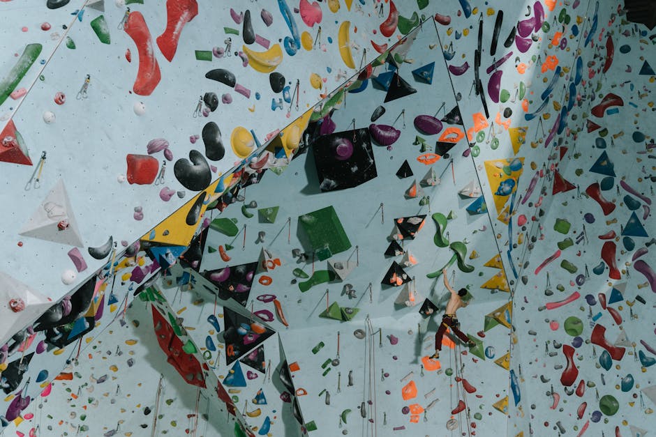 A man in casual clothing, wearing a black t-shirt and beige shorts, is loading a large white kitchen cabinet into the rear of a black moving van parked on a street. The van's back doors are open, revealing the interior where other packed boxes and packing materials are stored. The cabinet is being lifted from the ground and positioned inside the vehicle, with a hand gripping the side for stability. The surrounding environment includes a sidewalk with a potted shrub, and the scene appears to be part of a home relocation or furniture transport process operated by Man with Van Finsbury, as indicated by the company's branding on the van.