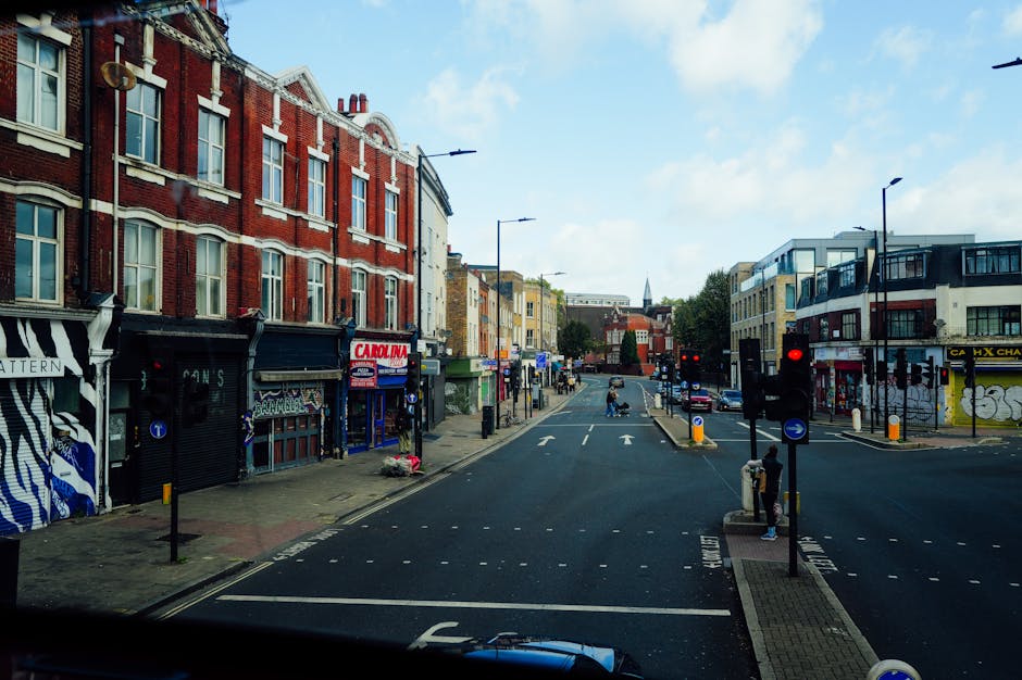 A street scene on Pentonville Road in Finsbury, showing a wide road with multiple lanes for vehicles and a central pedestrian island with a crossing. On the left side, there is a building with a brick
