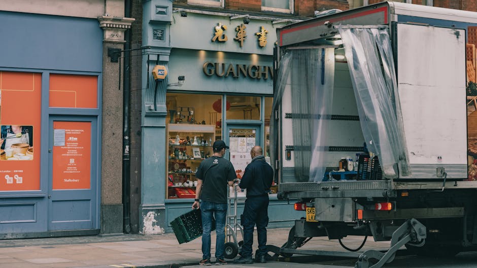 Two men with dark hair and dark jackets are standing on a sidewalk outside a storefront with an Asian-inspired sign reading 'GUANG' and Chinese characters above. One man is holding a small dolly, while the other appears to be giving instructions or discussing the move. A large, open-sided moving truck with a partially transparent plastic curtain on the side is parked nearby, with the rear loading area visible. Inside the truck, shelves hold various small boxes and items, with additional packing materials and equipment visible around the workspace. The building exterior includes blue and grey painted elements, with orange signage and posters on the adjacent wall. This scene depicts a home relocation process involving furniture and goods being prepared for transportation, coordinated by a professional removals company, evidenced by the presence of packing materials, transport equipment, and the loading process.
