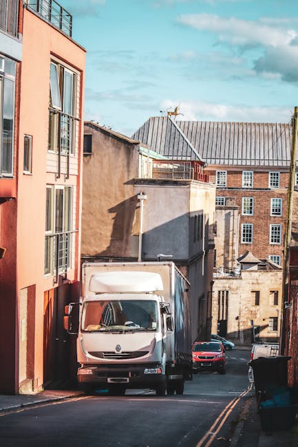 A white delivery van is parked on a narrow street, slightly inclined, surrounded by multi-storey residential buildings with various facades including brick, stucco, and painted surfaces. The van is positioned close to the building on the left, with its front facing the top of the image, and appears to be loading or unloading based on nearby activity. Behind the van, a pink car is parked along the curb, and another vehicle is partially visible further down the street. The street features double yellow lines, and a black wheelie bin is placed on the pavement near the curb on the right side. The buildings have multiple windows, some with small balconies, and a rooftop with a weather vane and a satellite dish can be seen in the background. The scene is illuminated by natural daylight, casting distinct shadows, and the overall setting reflects a typical residential area suitable for house removals and furniture transport managed by [COMPANY_NAME].