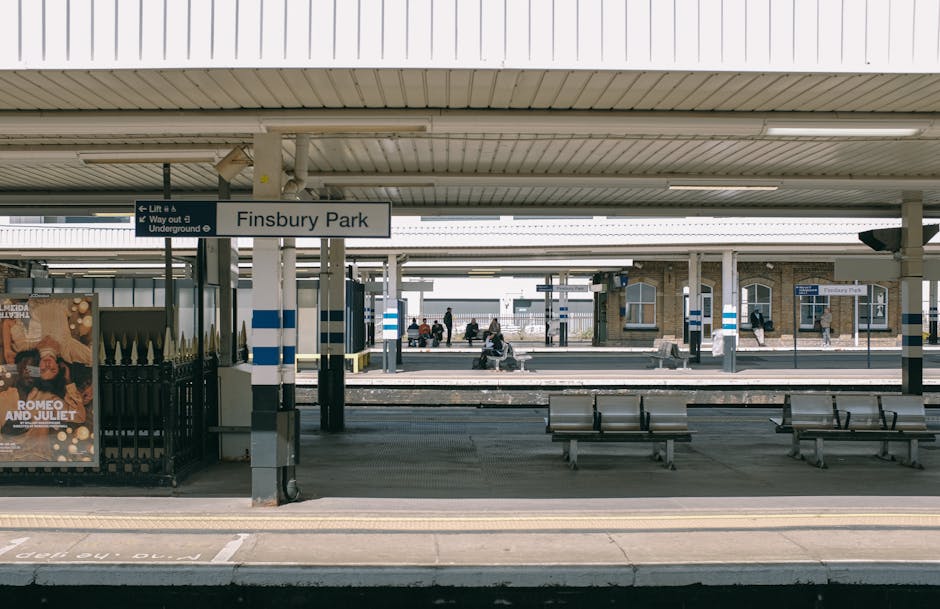 The image shows an outdoor view of Finsbury Park railway station platform, with a covered waiting area featuring metallic benches and a sign indicating the station name, 'Finsbury Park.' Several passengers are seated on the benches or standing nearby, while others are walking along the platform. To the left, there is a poster advertising 'Romeo and Juliet,' and in the background, additional platform areas extend with more people visible in the distance. The station has support columns painted with blue and white stripes, and overhead, there is a corrugated metal roof providing shelter. The scene is captured in daylight with natural lighting. This setting illustrates the typical environment where house removals or furniture transport might be coordinated, reflecting the importance of organized loading and unloading processes when a property, such as a Finsbury flat without lift access, involves moving heavy or bulky items to or from transportation vehicles managed by companies like Man with Van Finsbury.