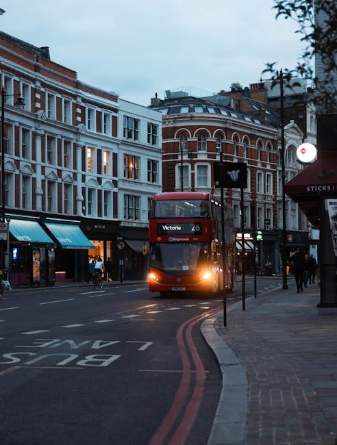 A red double-decker bus with the route number '26' and destination 'Victoria' displayed on the front is approaching a bus stop on a city street in Finsbury. The street features a mix of historic and modern buildings, with shopfronts and awnings lining the pavement. Pedestrians are walking along the sidewalk, some waiting near a bus stop sign. To the right, a red canopy over a shop entrance is visible, and street lighting begins to turn on as the sky appears overcast and slightly darkening, indicating early evening. The road has double red lines along the curb, and a 'BUS STOP' marking is painted on the asphalt. The scene captures the urban environment associated with city transport and local commerce, relevant to house removals and moving logistics in Finsbury, with Man with Van Finsbury's operational context subtly implied through the busy streetscape and transportation elements.
